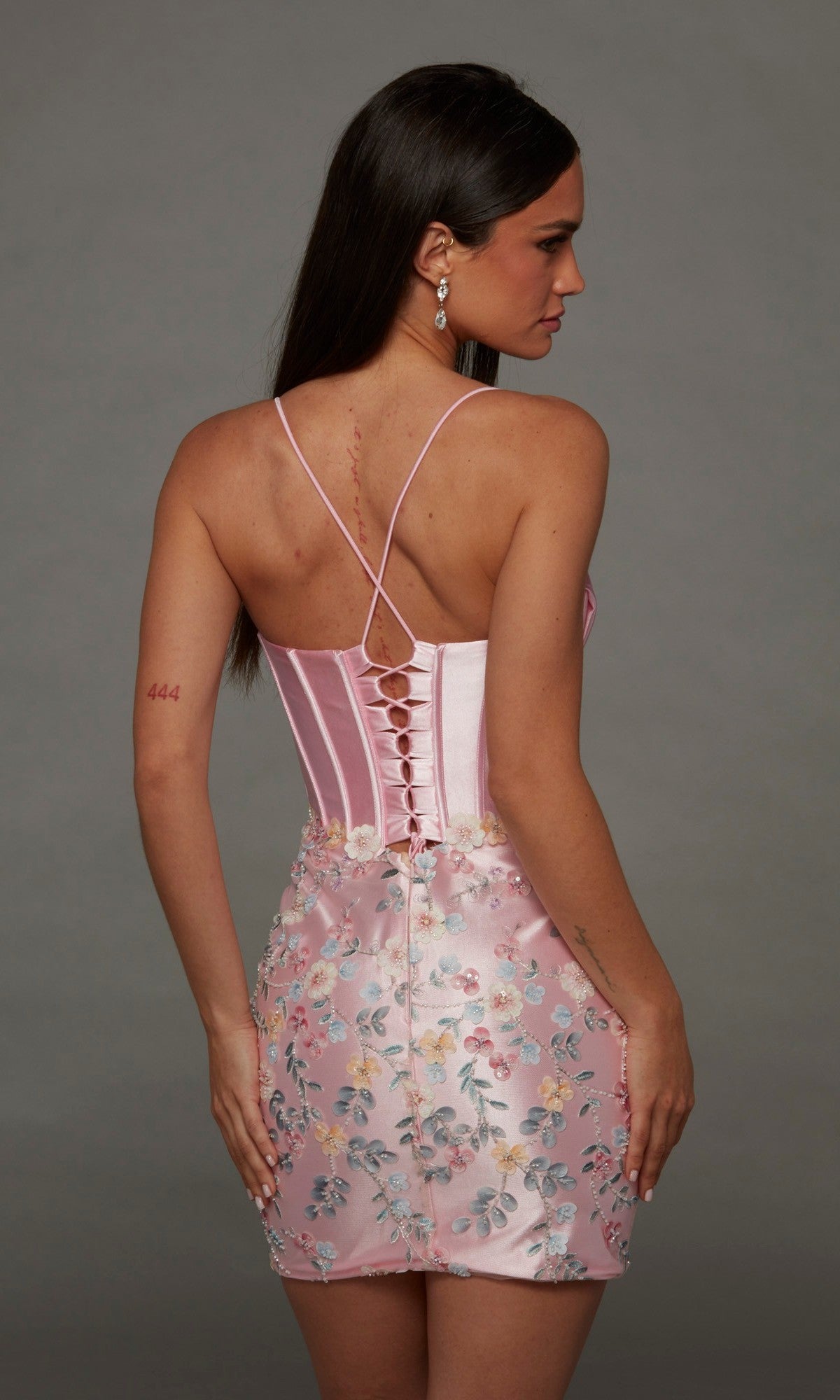 A woman is seen from behind wearing the Alyce 40140 Short Homecoming Dress, which features pink floral embroidery and a lace-up back. Her brown hair falls loose, she wears earrings, and a tattoo is visible on her upper left arm.